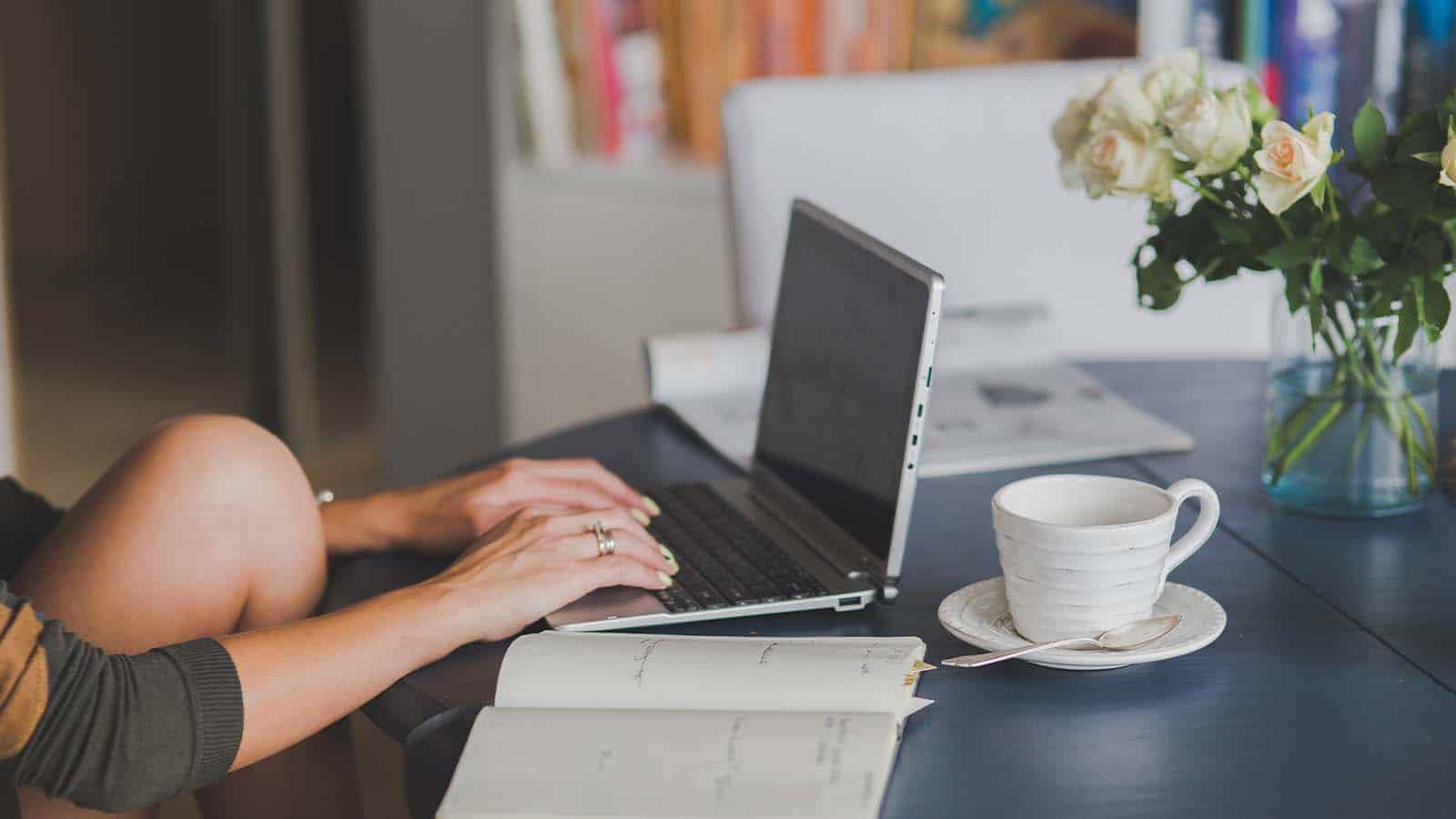 Person typing on a laptop on a table with a cup of coffee and a notebook