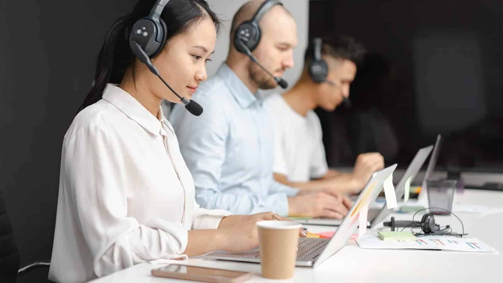 A person wearing headphones is intently listening to their computer while seated at their desk in an indoor workplace.