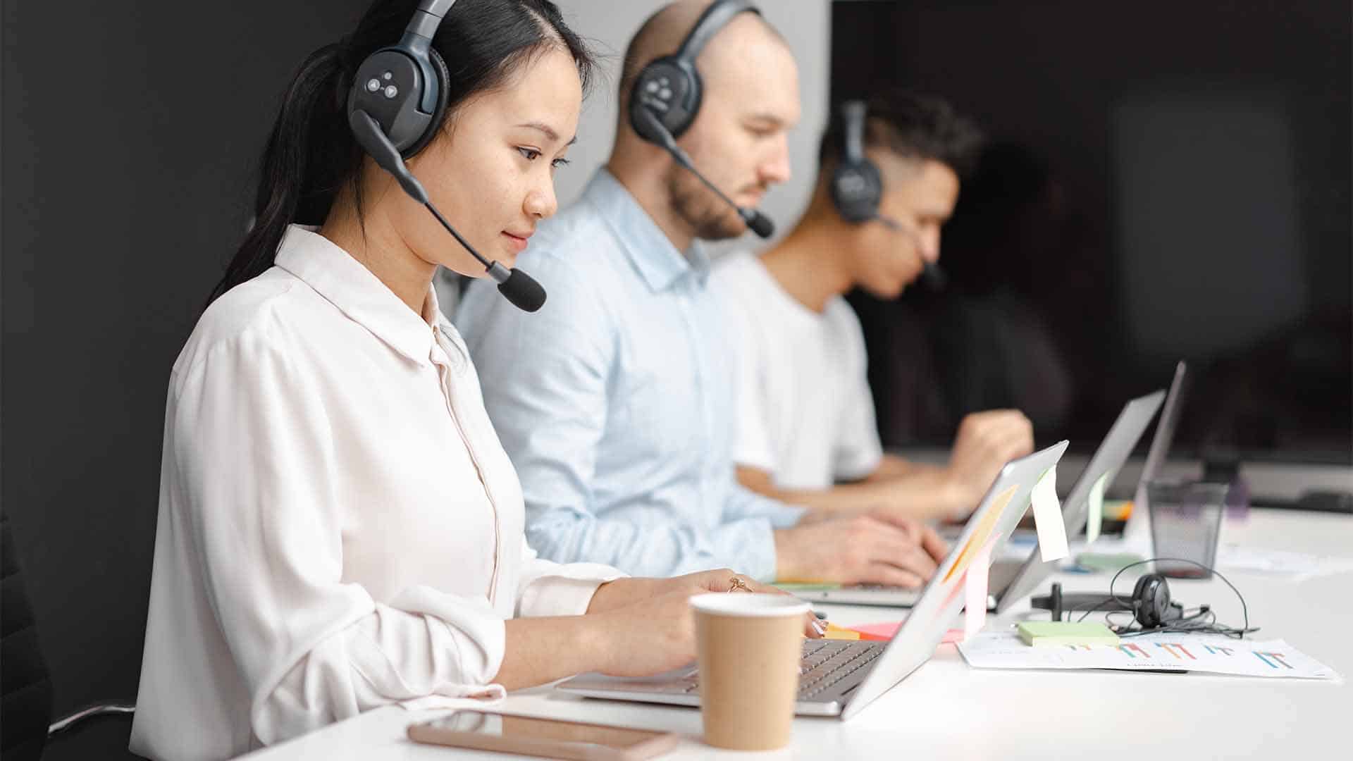 A person wearing headphones is intently listening to their computer while seated at their desk in an indoor workplace.