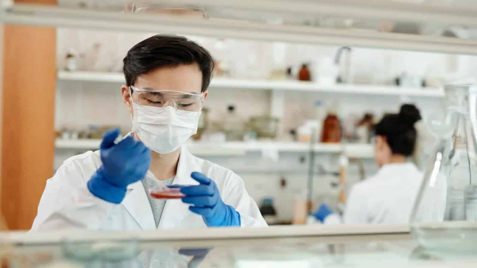 A medical technologist wearing a white coat and medical gloves is using laboratory and medical equipment to conduct research in a healthcare provider's clinic.