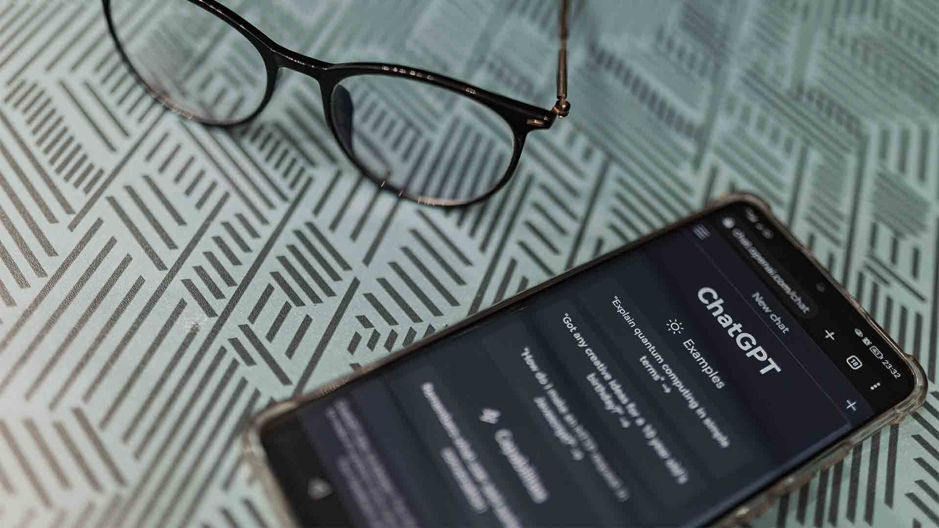 A smartphone with the ChatGPT app open lies on a patterned surface next to a pair of glasses, suggesting a work or study scenario.
