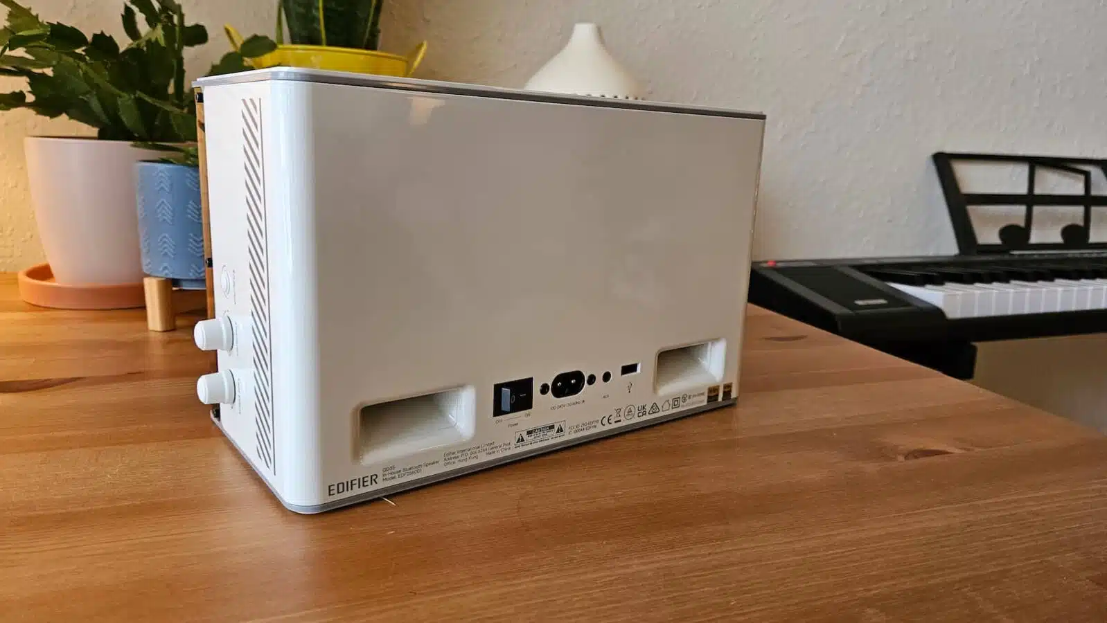 This image shows the back of a white Edifier speaker on a wooden table, featuring power and audio input ports, with plants and piano in background.