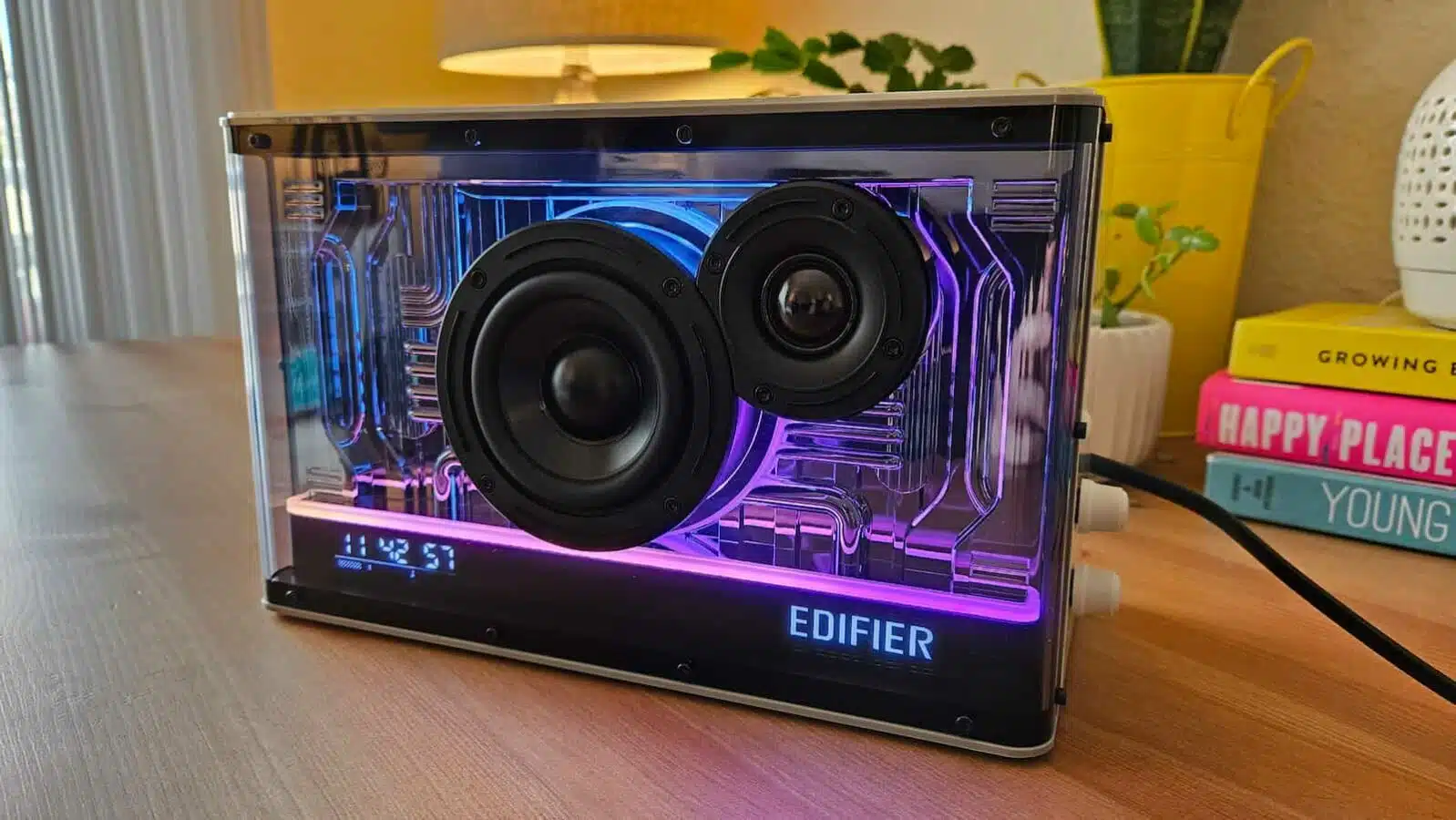 An Edifier transparent speaker with illuminated purple and blue internals is displayed on a wooden surface, with books and decor in the background.