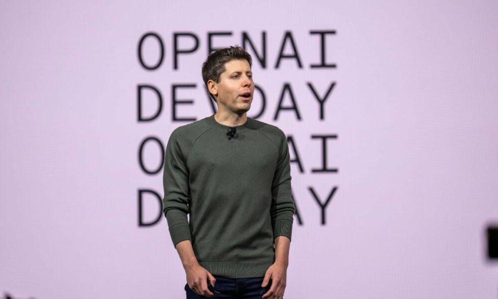 An individual is standing on stage with "OPENAI DEMO DAY" in the background, wearing a dark green sweater and dark pants, presenting to an audience.