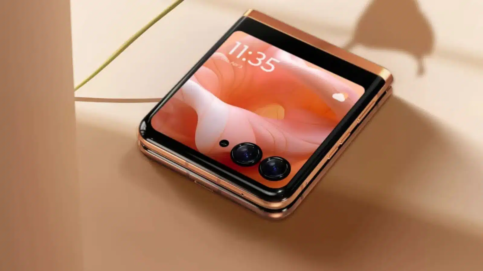 The image shows a modern foldable smartphone with a dual-camera setup lying on a beige surface, partially casting a shadow, displaying a colorful screen.