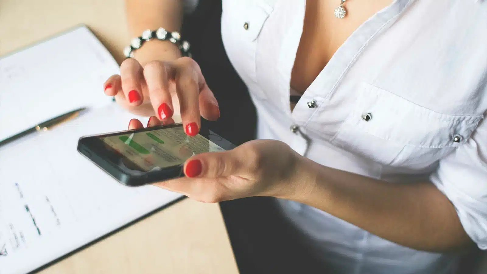 A person with red nail polish is using a smartphone over a desk with papers and a pen. The focus is on the hands and phone.