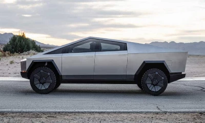 An angular, futuristic, silver pickup truck is parked on an asphalt road with a desert and mountains in the background at dusk.