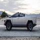 An angular, futuristic, silver pickup truck is parked on an asphalt road with a desert and mountains in the background at dusk.