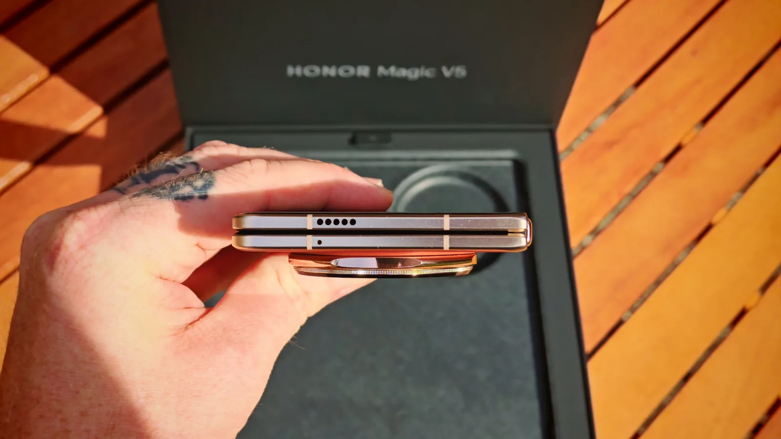 Flipped top view of a luxury Honor Magic V6 foldable smartphone showing sleek design and metallic finish, held by a person with tattoos, with the box and wooden surface background.
