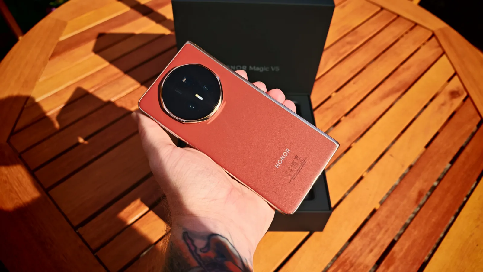 Vibrant Honor Magic V5 smartphone being held above a wooden table with a black box in the background, showcasing the sleek design and large camera module.