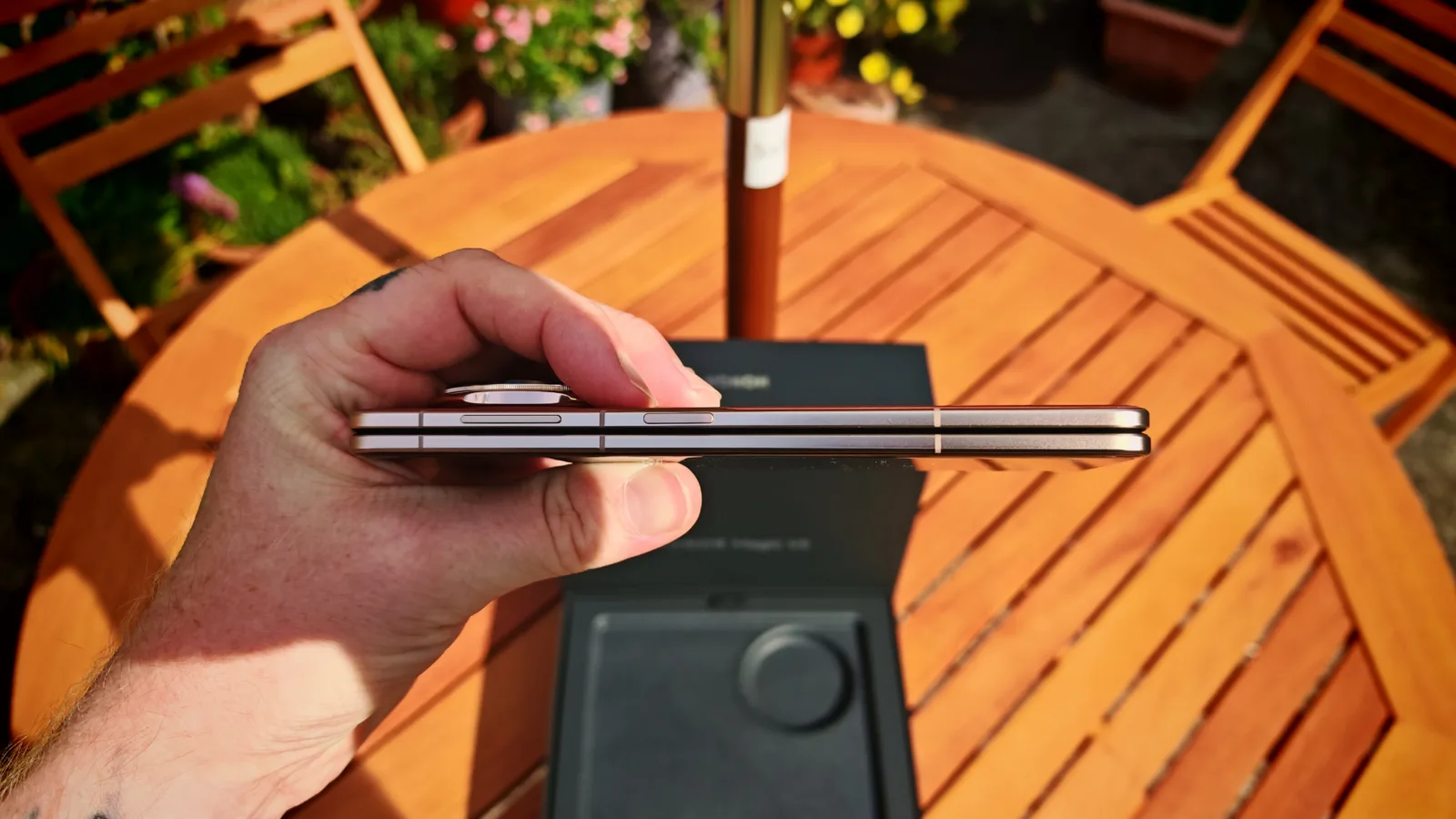 Slim modern smartphone with metallic edges, held in hand, showcasing its thin design, with outdoor wooden table and garden in background.