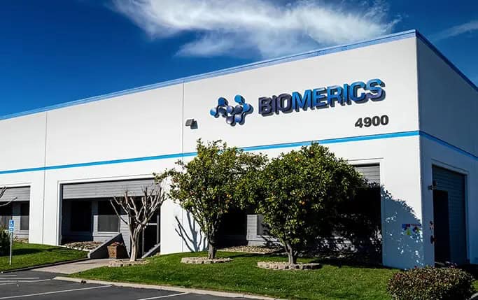 Biomerics biotech company exterior building with logo and company name, modern commercial facility in sunny weather, lush green trees, and well-maintained landscaping.