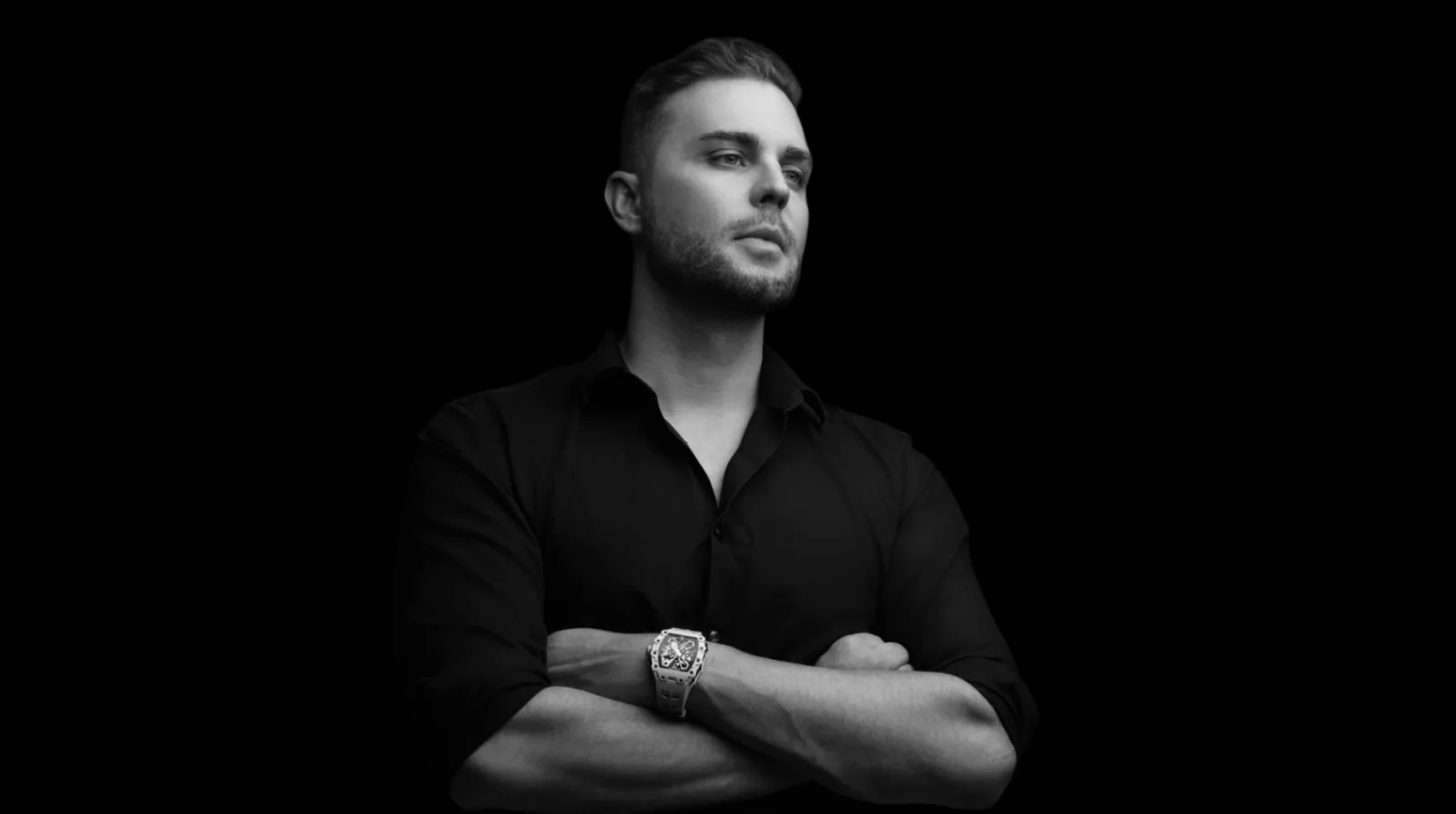 Male model with arms crossed, black shirt, and luxury watch, minimalist black background.