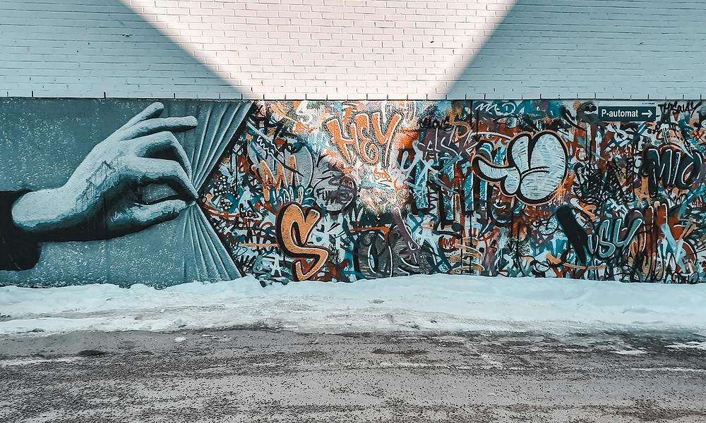 Hand pulling back a graffiti-covered wall mural featuring vibrant street art, graffiti tags, and illustrative designs, set against a brick building exterior with snow-covered ground.
