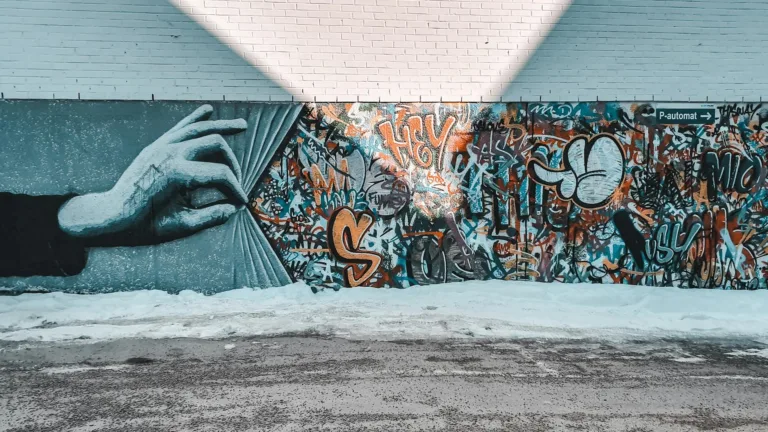 Hand pulling back a graffiti-covered wall mural featuring vibrant street art, graffiti tags, and illustrative designs, set against a brick building exterior with snow-covered ground.