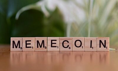 A close-up of wooden Scrabble tiles spelling "MEMECOIN" on a wooden surface with a soft-focus green leafy background, illustrating cryptocurrency memes and meme coins in the context of blockchain and digital finance.