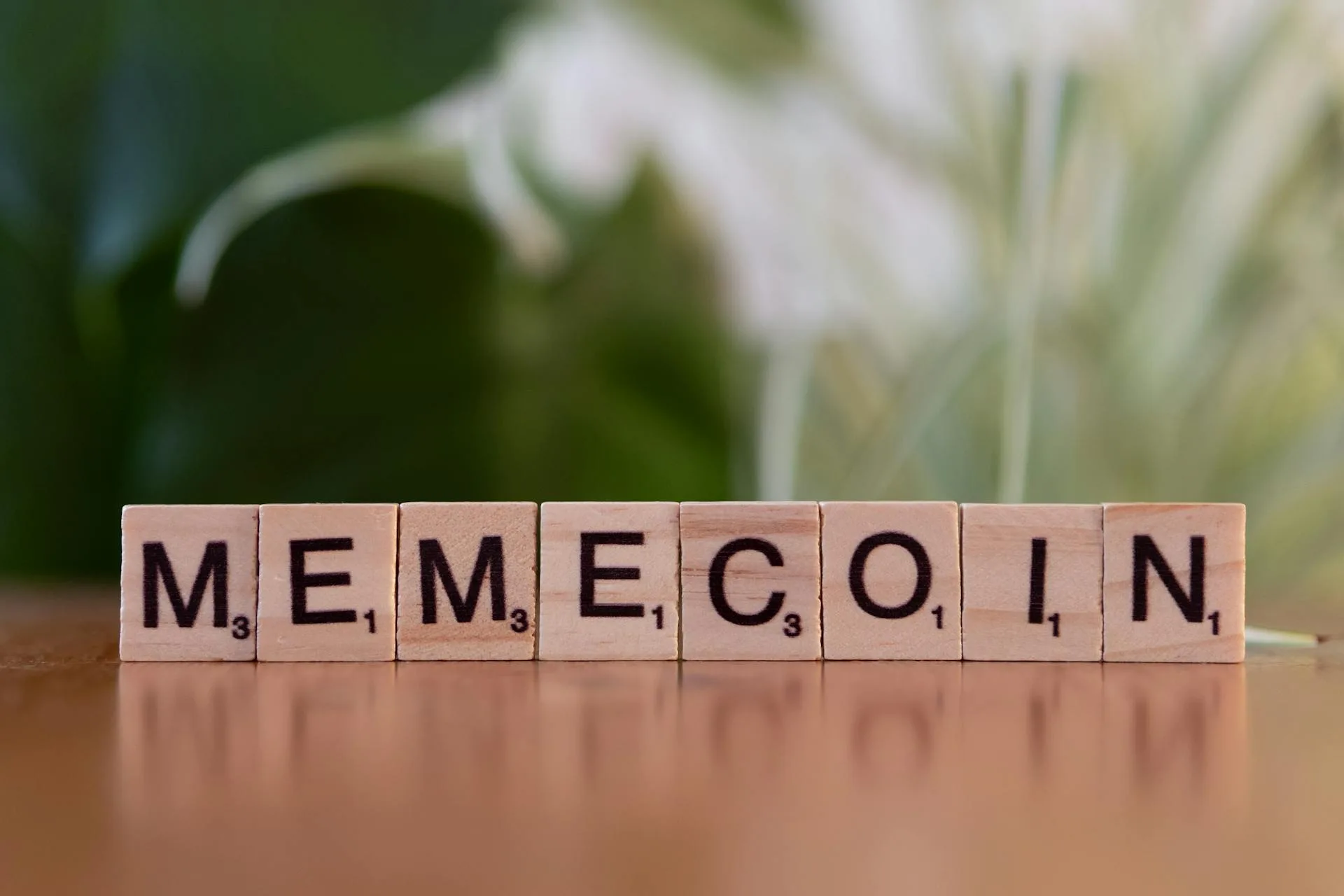 A close-up of wooden Scrabble tiles spelling "MEMECOIN" on a wooden surface with a soft-focus green leafy background, illustrating cryptocurrency memes and meme coins in the context of blockchain and digital finance.