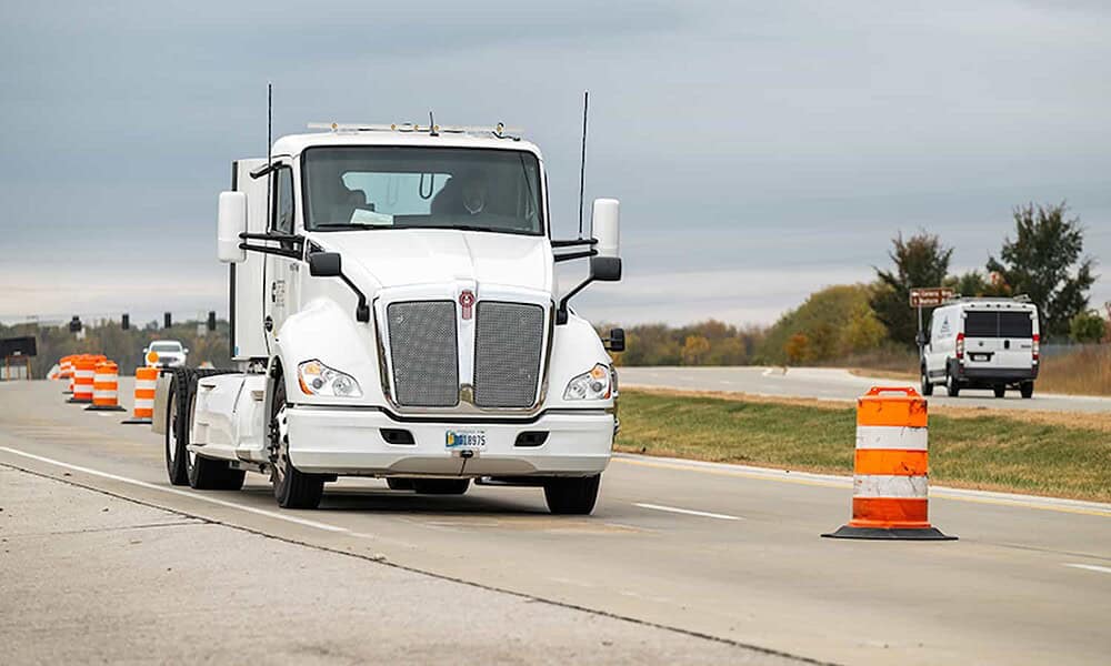 Large white semi-truck driving through road construction zone with orange safety barrels and traffic control devices.
