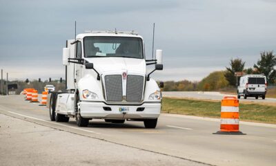 Large white semi-truck driving through road construction zone with orange safety barrels and traffic control devices.