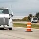 Large white semi-truck driving through road construction zone with orange safety barrels and traffic control devices.
