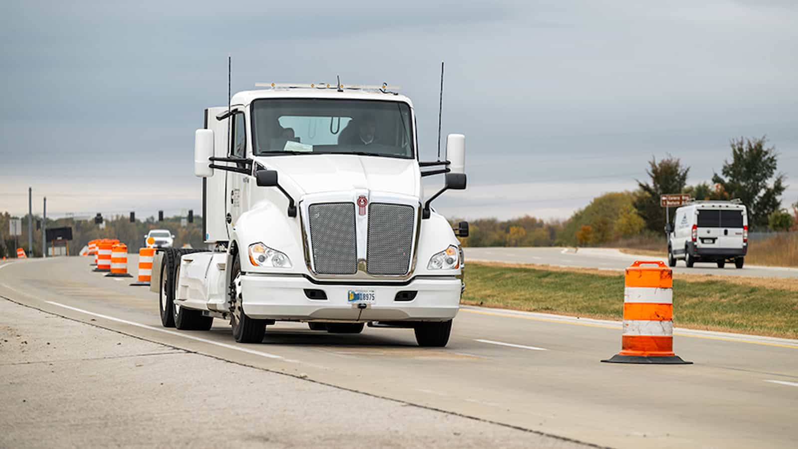 Large white semi-truck driving through road construction zone with orange safety barrels and traffic control devices.