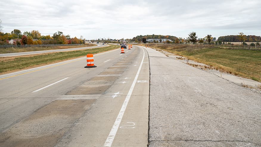 Construction cones and marked lanes on a highway under roadwork, with a rural landscape and cloudy sky in the background.