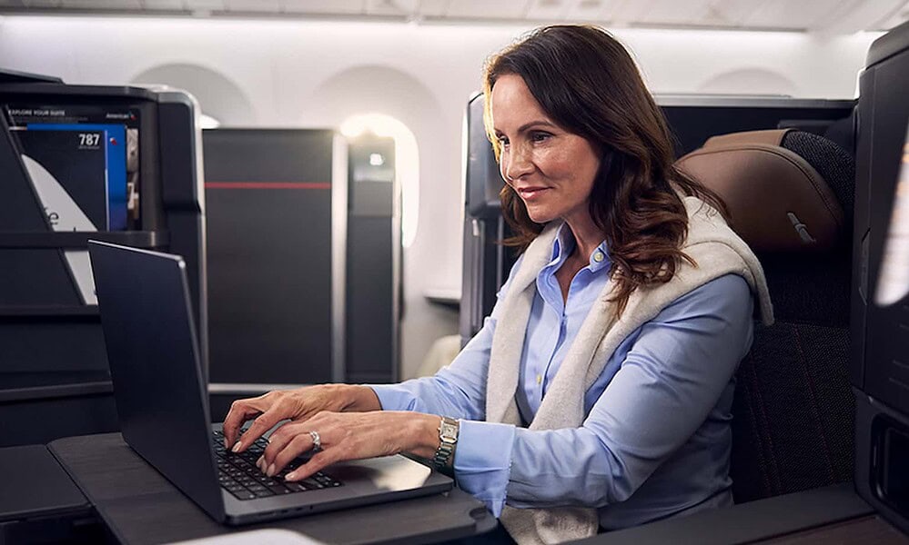 An airline passenger, a woman with brown hair, working on her laptop comfortably seated in an airplane's window seat during a flight. She is dressed in a light blue shirt with a gray vest, with her hands on the keyboard, surrounded by personal luggage and electronics, with sunlight filtering through the windows.
