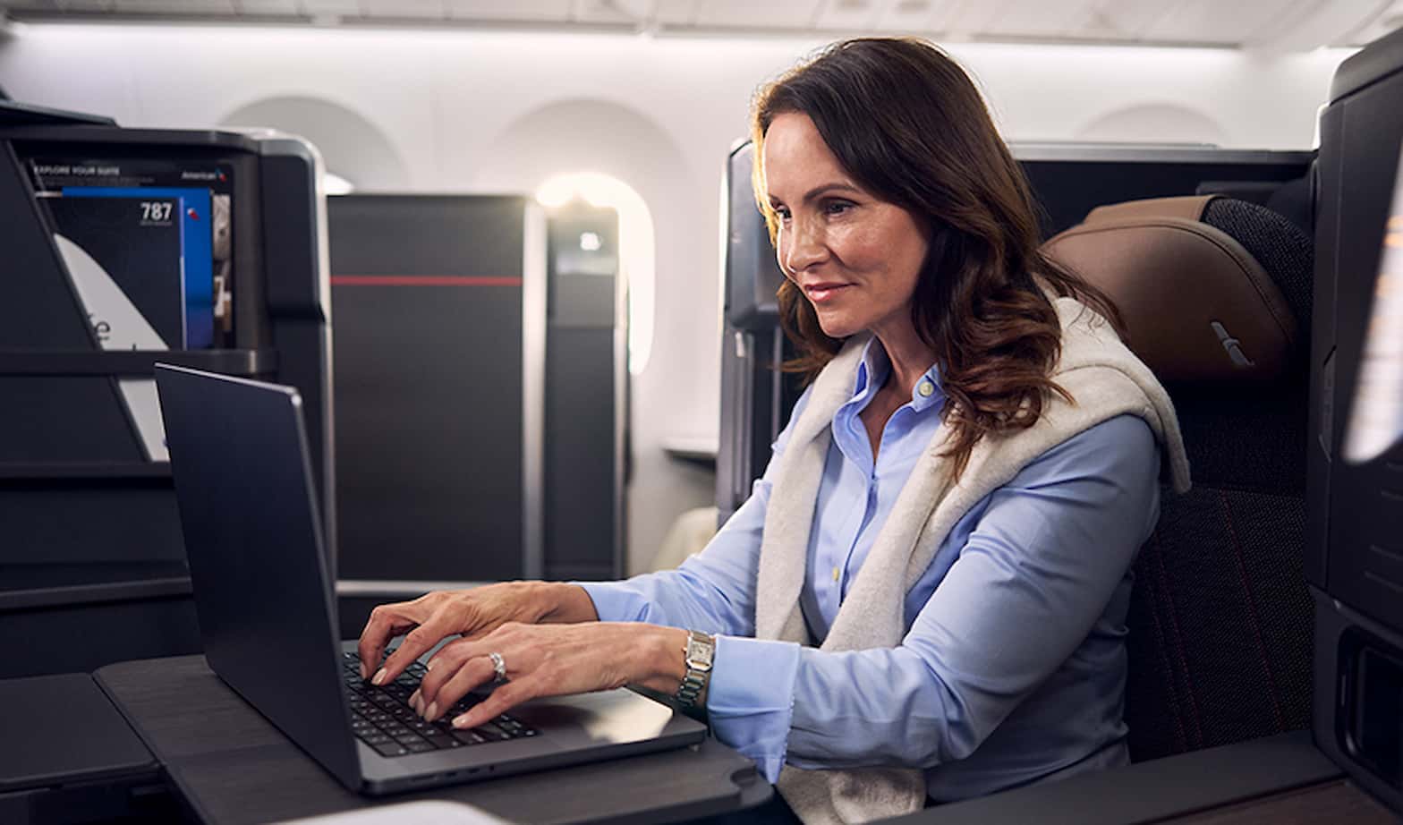 An airline passenger, a woman with brown hair, working on her laptop comfortably seated in an airplane's window seat during a flight. She is dressed in a light blue shirt with a gray vest, with her hands on the keyboard, surrounded by personal luggage and electronics, with sunlight filtering through the windows.