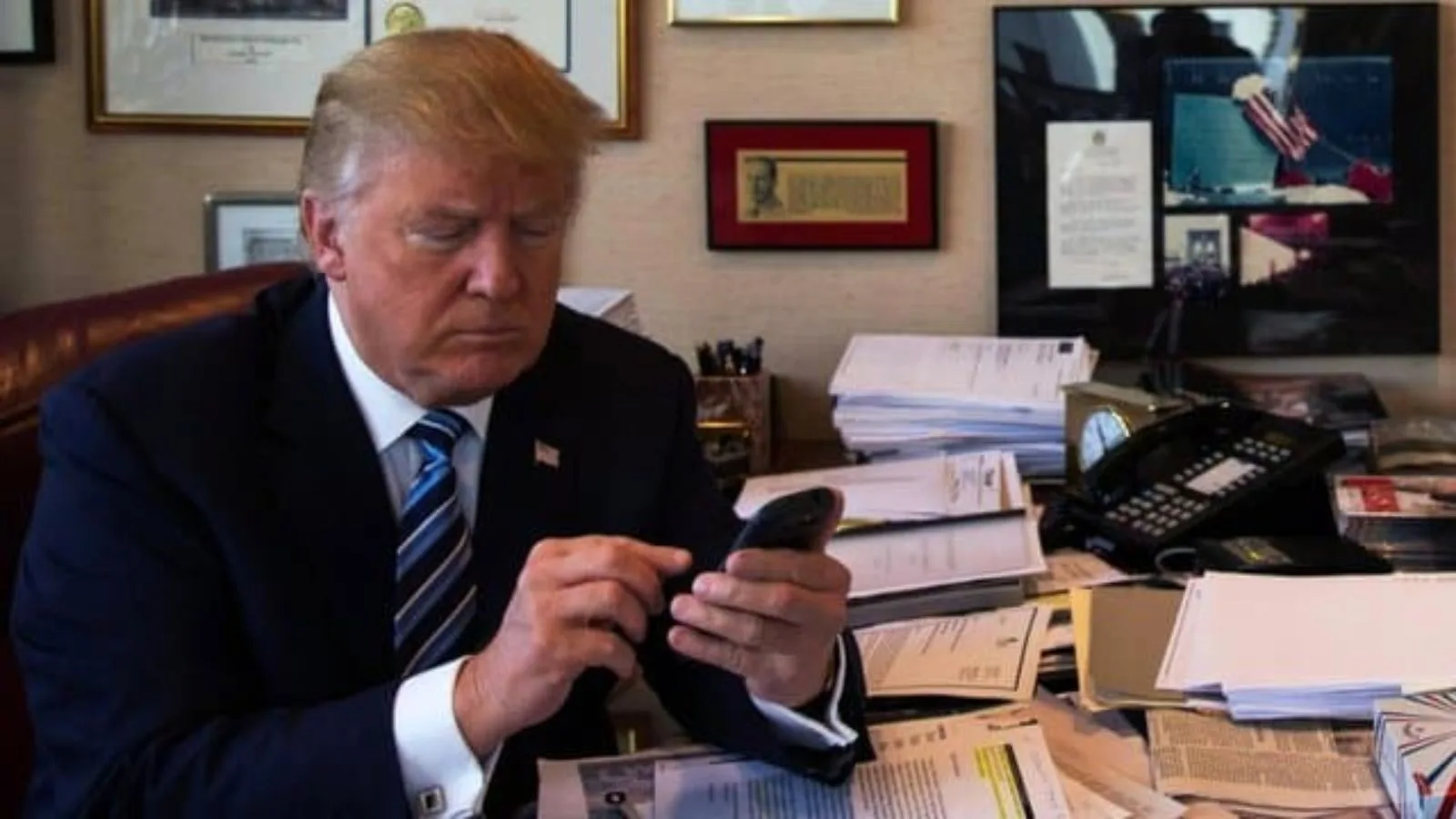 A political figure, possibly Donald Trump, focused on his mobile device amidst a busy work environment.